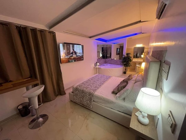Une chambre d’hôtel moderne, équipée d’un lit, d’une table de chevet avec une lampe, d’une télévision et d’une salle de bain visible à travers une porte en verre.