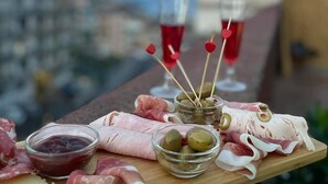 A charcuterie board with various meats, olives, and a glass of red wine on a balcony overlooking a cityscape.