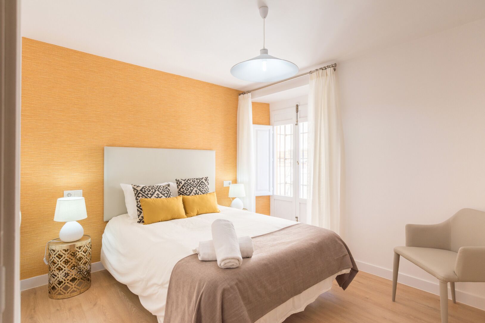 Een keurig opgemaakt slaapkamer met een bed, nachtlampjes, een stoel en een raam met gordijnen.