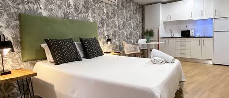 A hotel room with a bed, a desk, a chair, a small table with a plant, and a kitchenette.