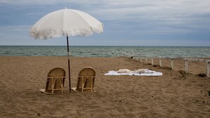 Una platja amb un para-sol blanc, dues cadires de vimet i una tovallola blanca sobre la sorra.