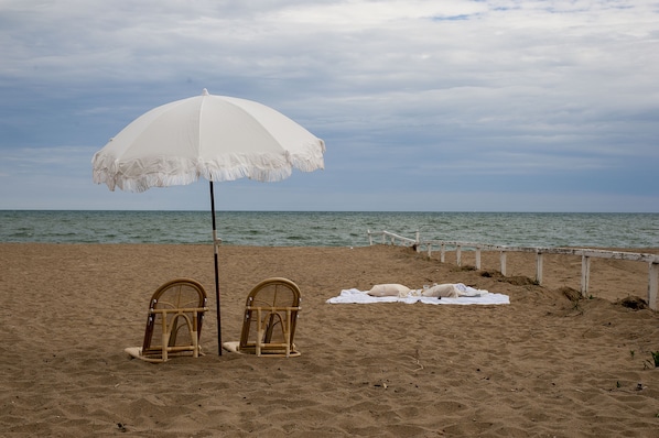 A beach with a white umbrella, two wicker chairs, and a white towel on the sand. - WaveCrest Cottage -KINGSVILLE - Premium Comfort (Kingsville)