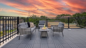 Eine Terrasss mit Sofa, Sesseln und Tisch mit Blick auf einen Parkplatz und Grünflächen.