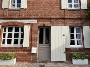 A brick building with white-framed windows and shutters, a central door, and two potted plants.