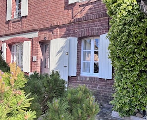 Un bâtiment en briques rouges avec des volets blancs, une enseigne portant le chiffre « 6 » et une haie verdoyante et luxuriante.