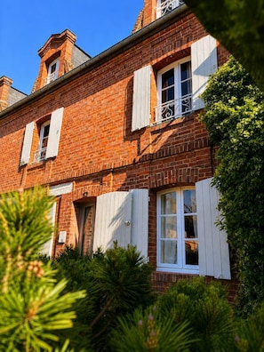 Un bâtiment en briques rouges avec des volets blancs, un balcon doté de balustrades en métal et, au premier plan, une végétation luxuriante.