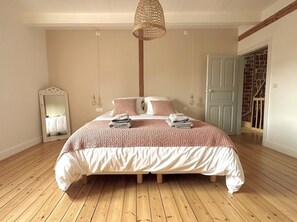 Une chambre avec un grand lit, un plancher en bois, un luminaire suspendu, un miroir et une porte donnant sur un escalier.