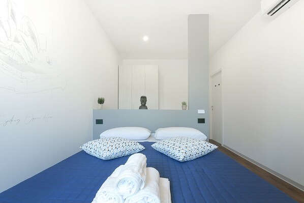 Quarto com cama, travesseiros, cobre-leito azul, espelho e parede com padrão geométrico.