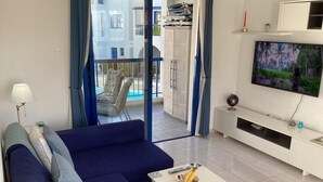 A modern living room with a blue sofa, a white coffee table with a magazine, a TV on the wall, and a balcony with a view of buildings. -  Pool view apartment, walk to the beach and harbour (Paphos)