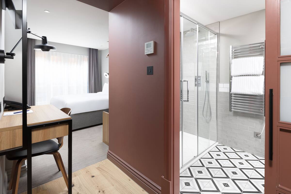 Ett hotellrum med ett skrivbord i trä, en stol, en säng, en dusch med glasväggar och ett badrumsgolv med kakel.