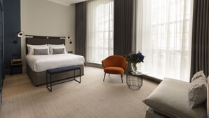 A modern hotel room with a large bed, a bench, an orange armchair, and a view of the city through large windows.