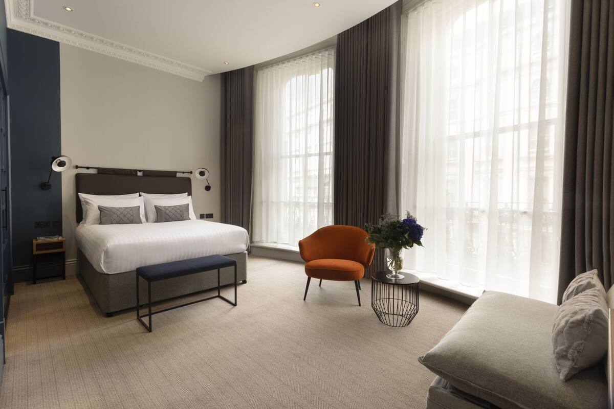 Ett modernt hotellrum med en stor säng, en bänk, en orange fåtölj och utsikt över staden genom stora fönster.