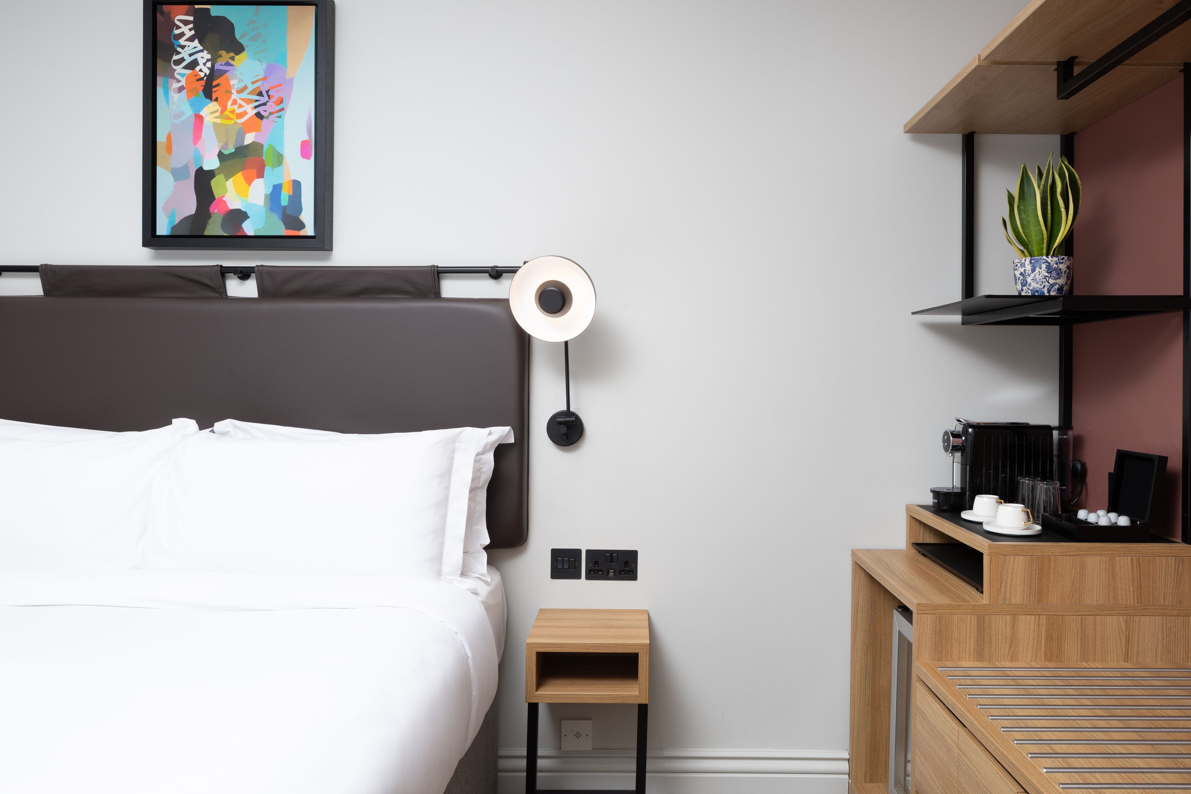 A modern hotel room with a bed, bedside table, and a small wooden nightstand. There is a wall-mounted lamp, a framed abstract painting, and a shelf with a potted plant.
