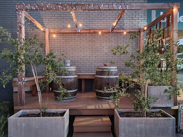 An outdoor seating area with wooden tables and chairs, surrounded by planters and barrels.