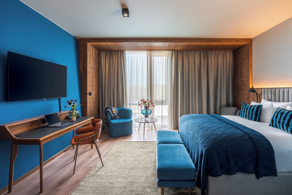 A modern hotel room with a large bed, a desk with a laptop, a TV, and a view of the outdoors through large windows.