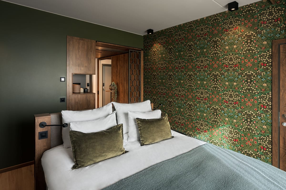 A neatly made bed with white linens and two green pillows, a wooden headboard, and a wall with a floral patterned wallpaper.