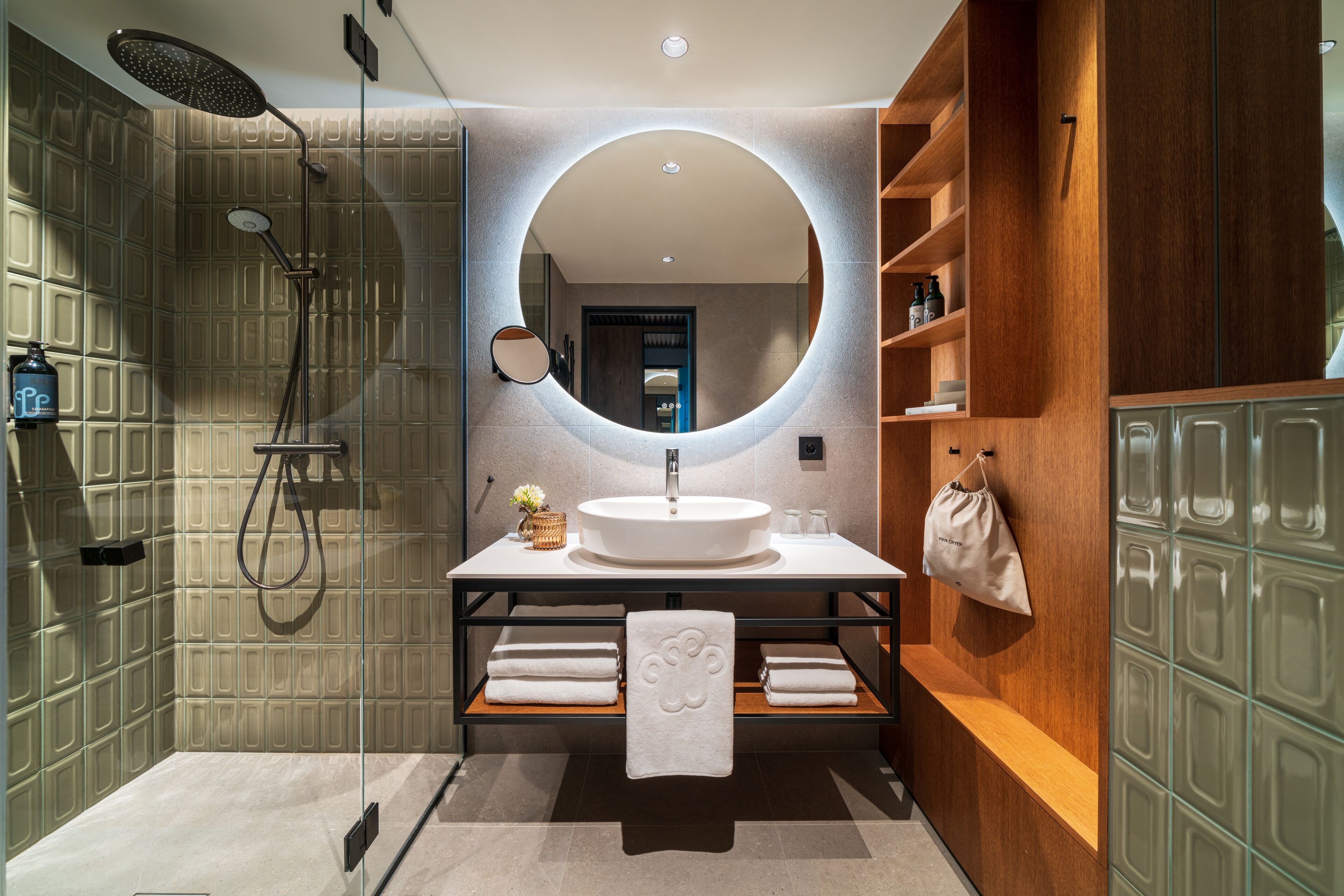 Un baño moderno con un espejo grande, un lavamanos redondo y una ducha. Se observan estantes de madera y un perchero para toallas.