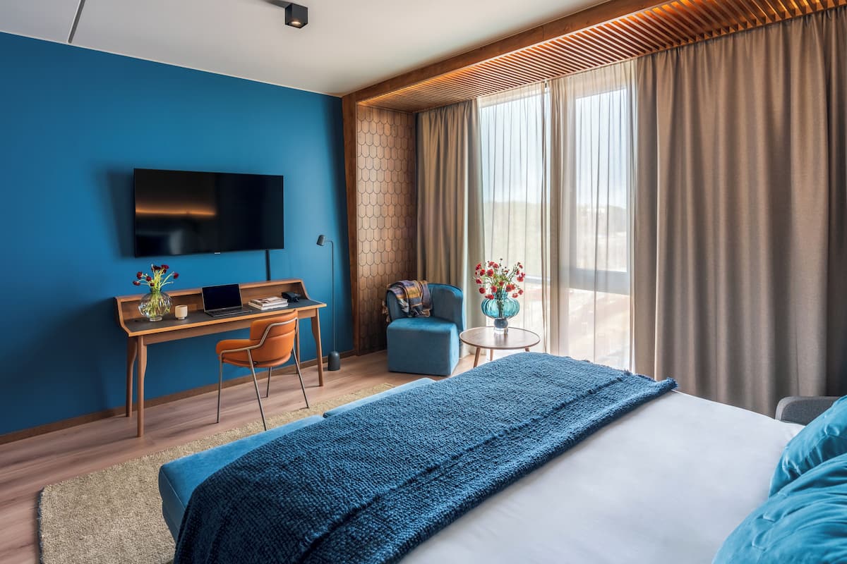 A modern hotel room with a large bed, a desk with a laptop, a TV, and a view of the outdoors.
