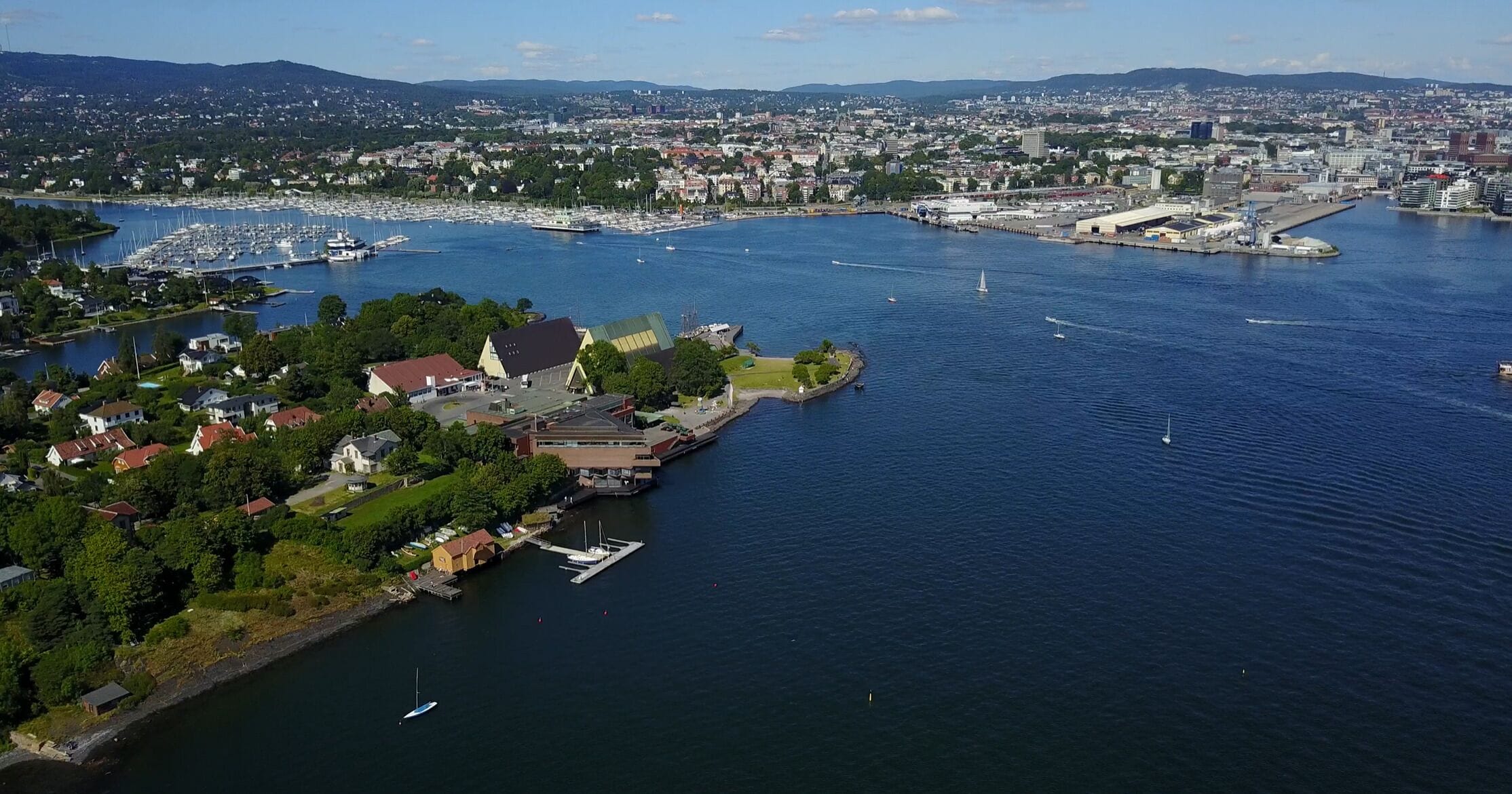 An aerial view of a coastal city with a marina, residential areas, and a prominent building with a green roof.