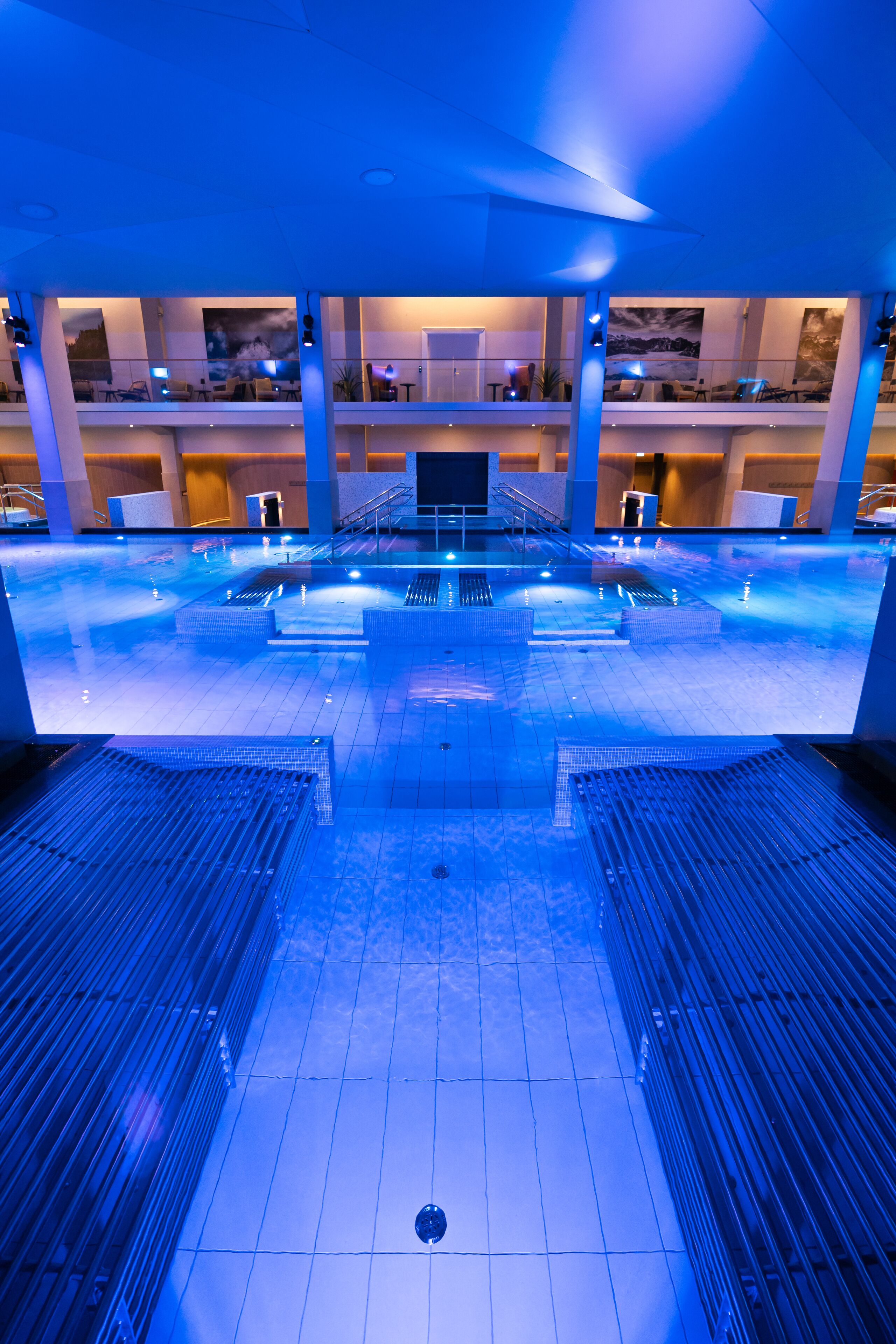 En stor indendørs swimmingpool med moderne design, omgivet af en flerniveaukonstruktion med siddepladser og udsigt over poolen.