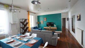 En moderne stue med en teal farvet væg, et spiseområde med hvide tallerkener og glas, samt en grå sofa.