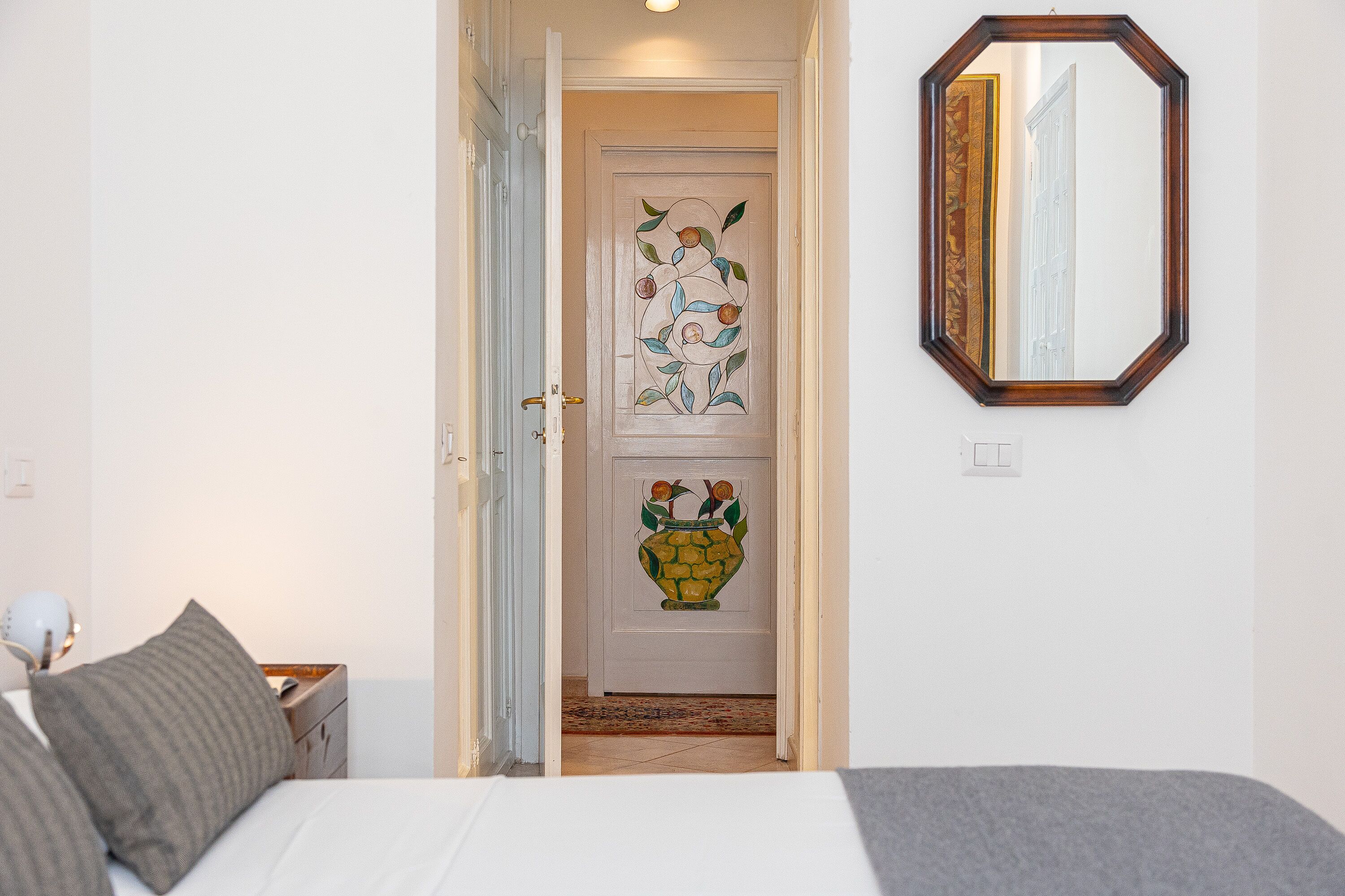A bedroom with a bed, a nightstand, a lamp, a mirror, and a door with a stained glass design.