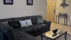 A living room with a black leather sofa, a glass coffee table, and a welcome sign on the wall. - Pool home in Phoenix Retreat close to everything! (Phoenix)