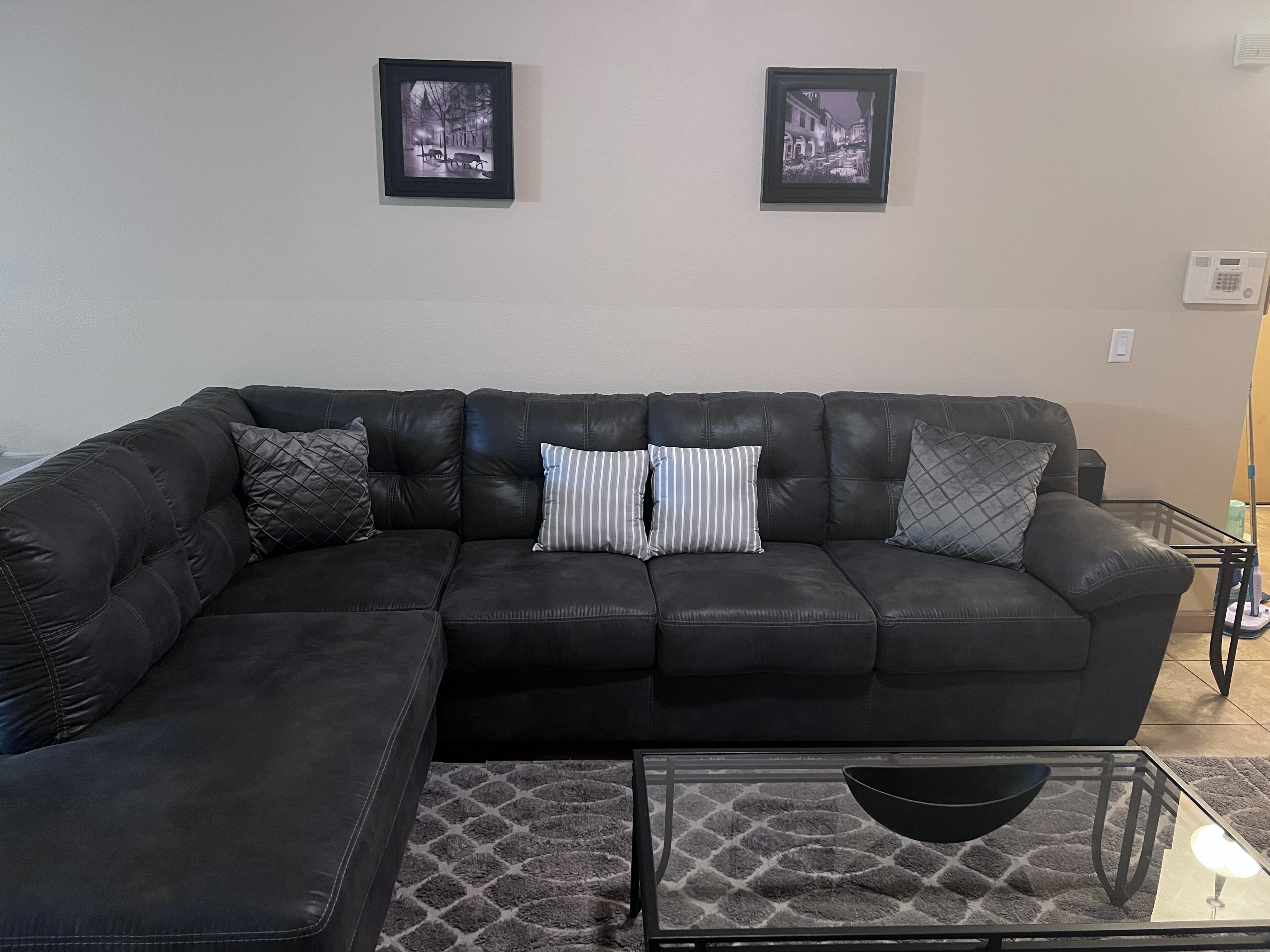 A living room with a black leather sectional sofa, two framed pictures on the wall, and a glass coffee table.