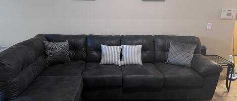 A living room with a black leather sectional sofa, two framed pictures on the wall, and a glass coffee table.