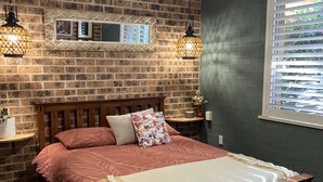 Ein Schlafzimmer mit Backsteinwand, einem hölzernen Bett, zwei Wandleuchten, einem Deckenventilator und einem Fenster mit Jalousien.