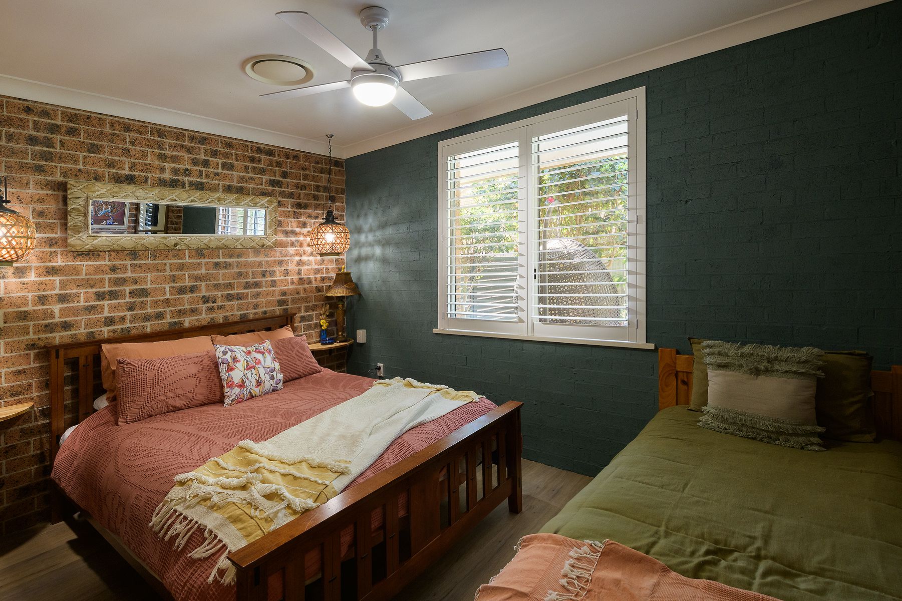 Een slaapkamer met een bakstenen muur, een bed met houten hoofdbord, een raam met zonwering en een plafondventilator.