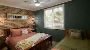 Een slaapkamer met een bakstenen muur, een bed met houten hoofdbord, een raam met zonwering en een plafondventilator.