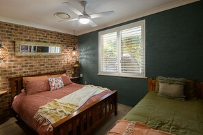 A bedroom with a brick wall, a bed with a wooden headboard, a window with blinds, and a ceiling fan. - Salty Crew villa , Central spot , Pet friendly ,private garden oasis secure yard (Hawks Nest)