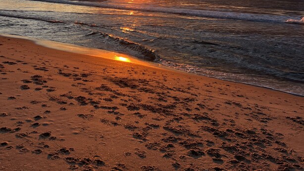 Ein Strand mit Fußspuren, die zum Wasser führen, Wellen und einem Sonnenuntergang.