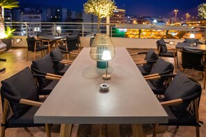 A rooftop terrace with a long table and chairs, a decorative lamp, and a view of the cityscape at night. - MY WAY HOTEL & EVENTS (Patras)