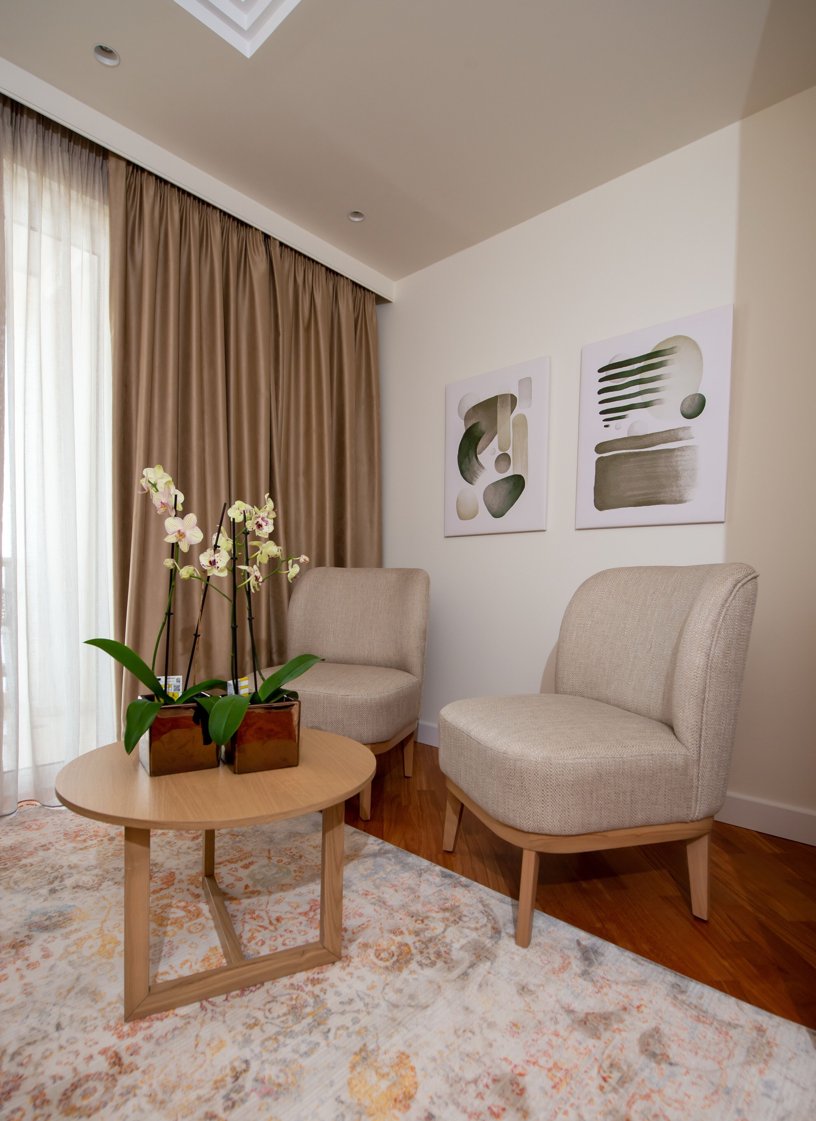 A living room with two beige armchairs, a round wooden coffee table, and a floral-patterned rug. There are two orchid plants in a wooden planter on the table. The room has beige curtains, a ceiling light, and two abstract art pieces on the wall.