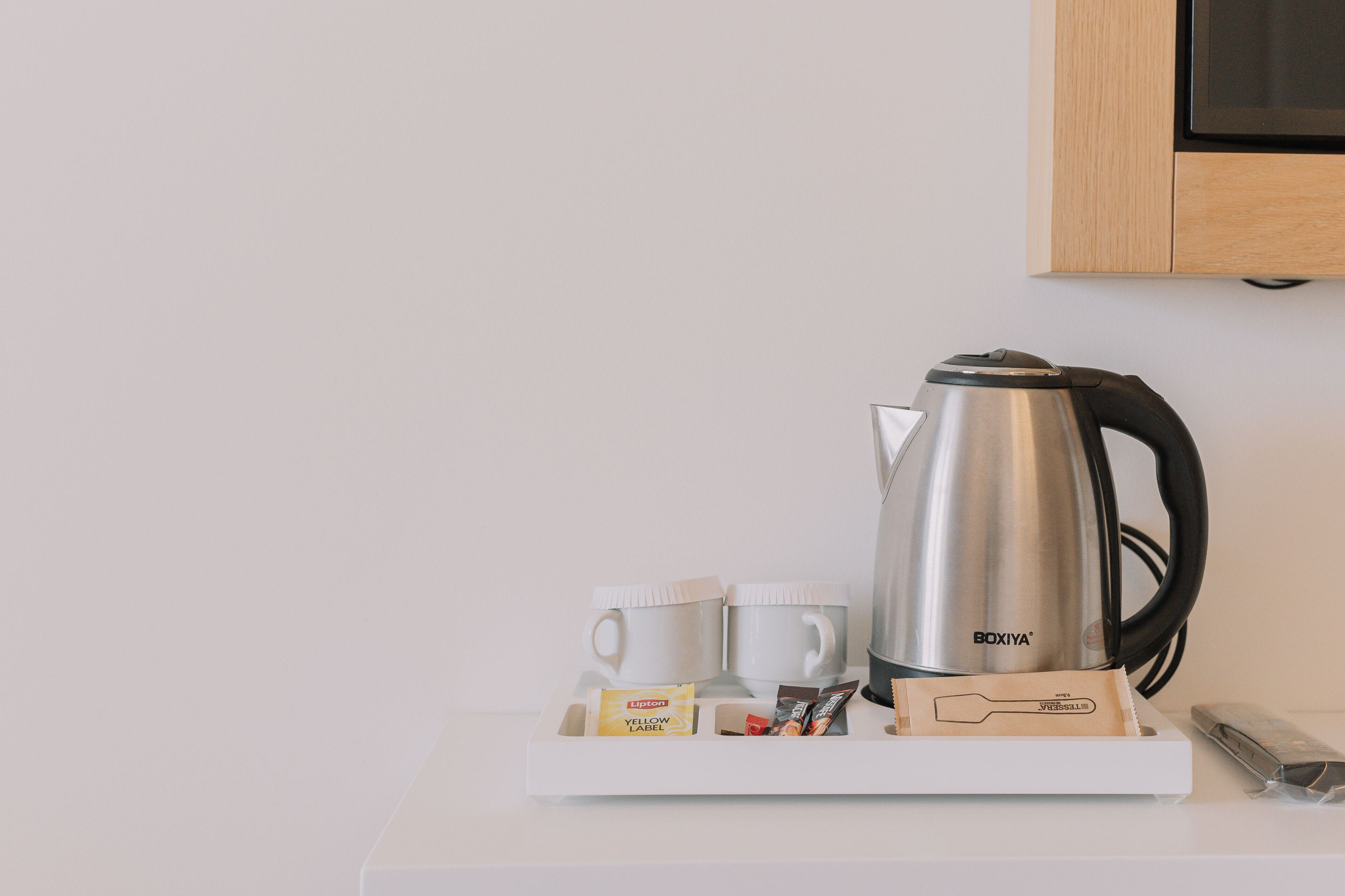 Una cafetera de acero inoxidable, dos tazas blancas, una bandeja con bolsitas de té Lipton y un control remoto sobre una mesa blanca.