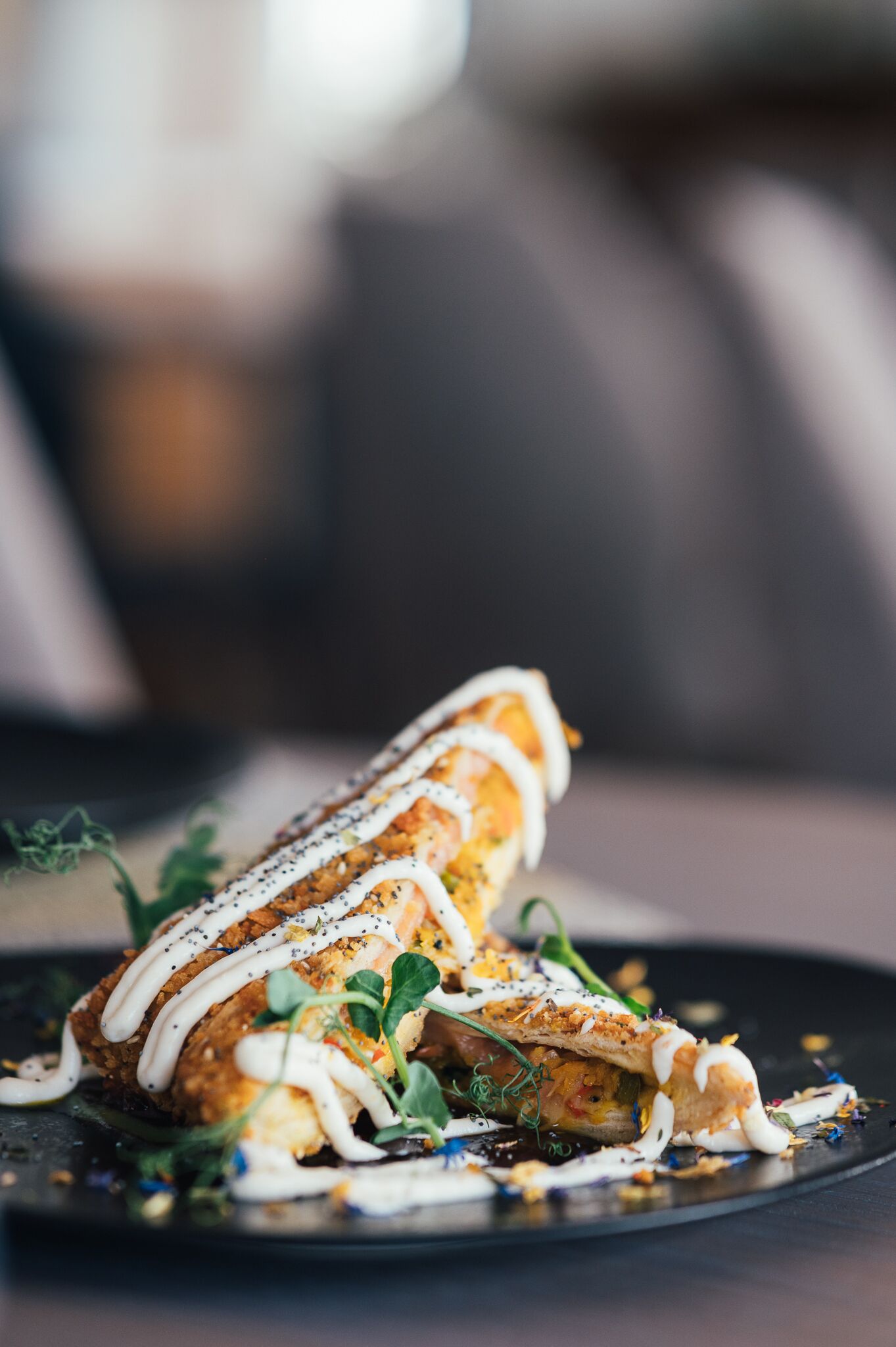 A dish with a crispy exterior, drizzled with white sauce, garnished with herbs, and served on a dark plate.