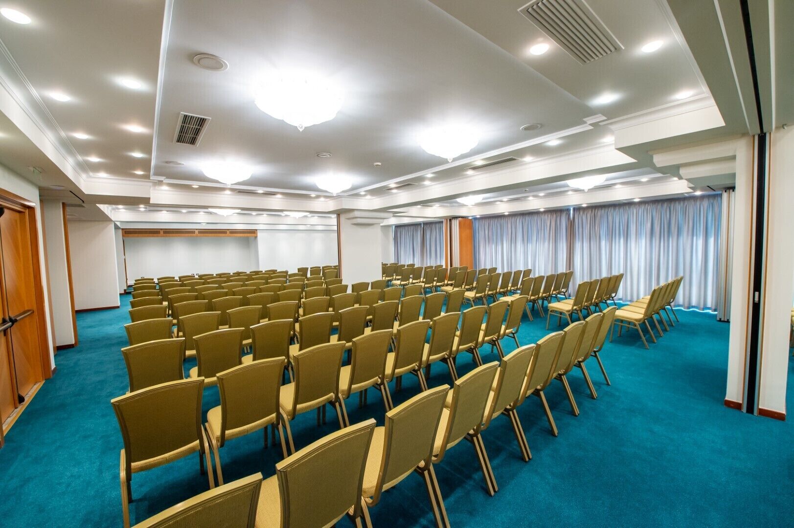 Un salón de conferencias con filas de sillas, alfombra azul y techo blanco con iluminación empotrada.