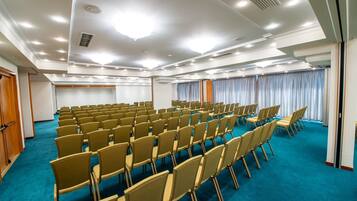 Un salón de conferencias con filas de sillas, alfombra azul y techo blanco con iluminación empotrada.
