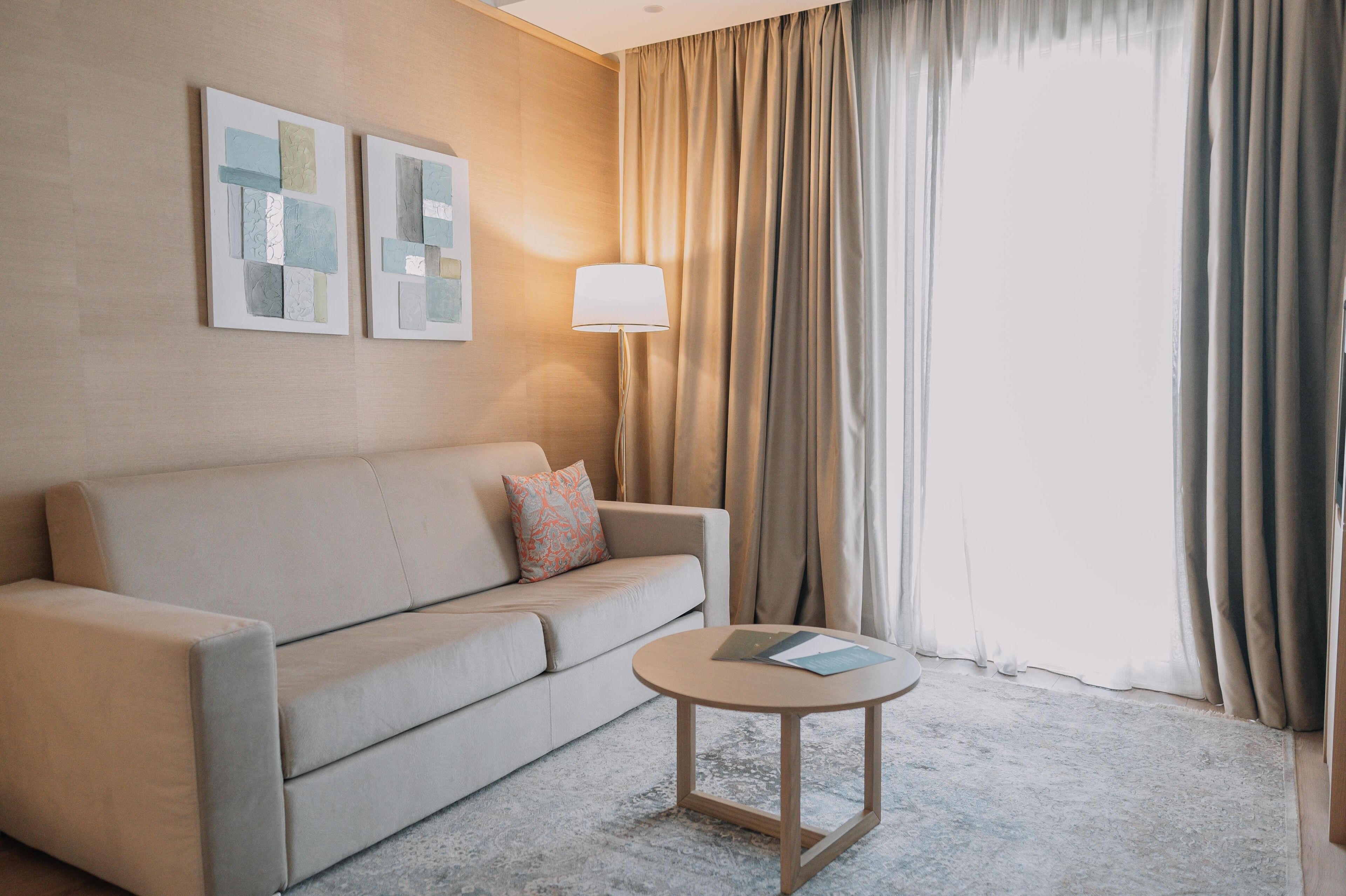 A hotel room with a beige sofa, a round wooden table, and a large window with beige curtains.