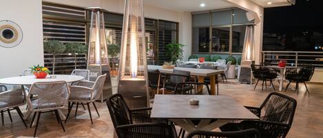 A modern outdoor dining area with round tables and black wicker chairs, surrounded by large windows and potted plants.