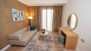 Una habitación de hotel moderna con un sofá color beige, una mesa redonda con un florero, un escritorio de madera y un espejo grande.
