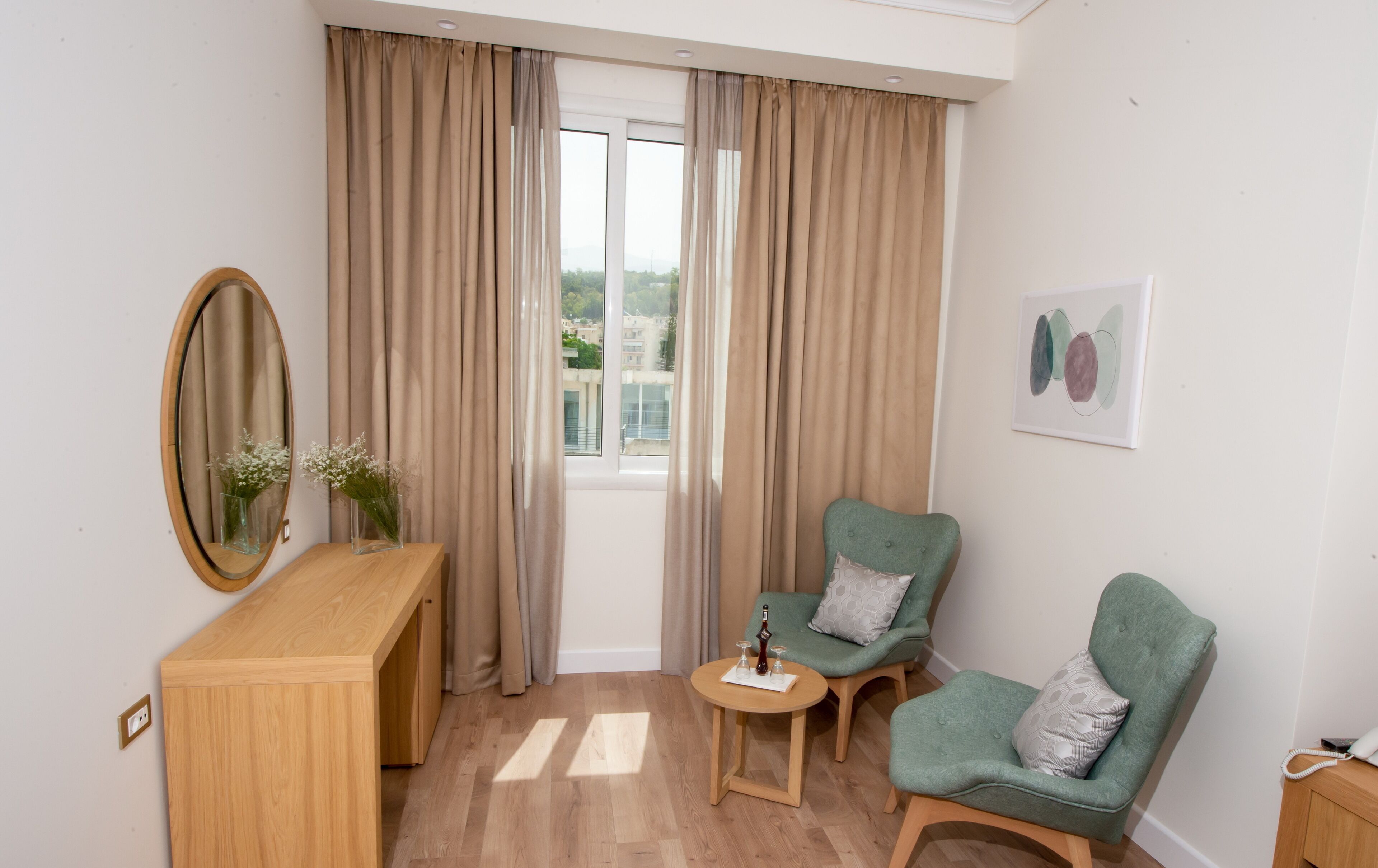 A room with a wooden desk, two green armchairs, a small round table, a mirror, beige curtains, and a window with a view of buildings.