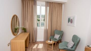 A room with a wooden desk, two green armchairs, a small round table, a mirror, beige curtains, and a window with a view of buildings.