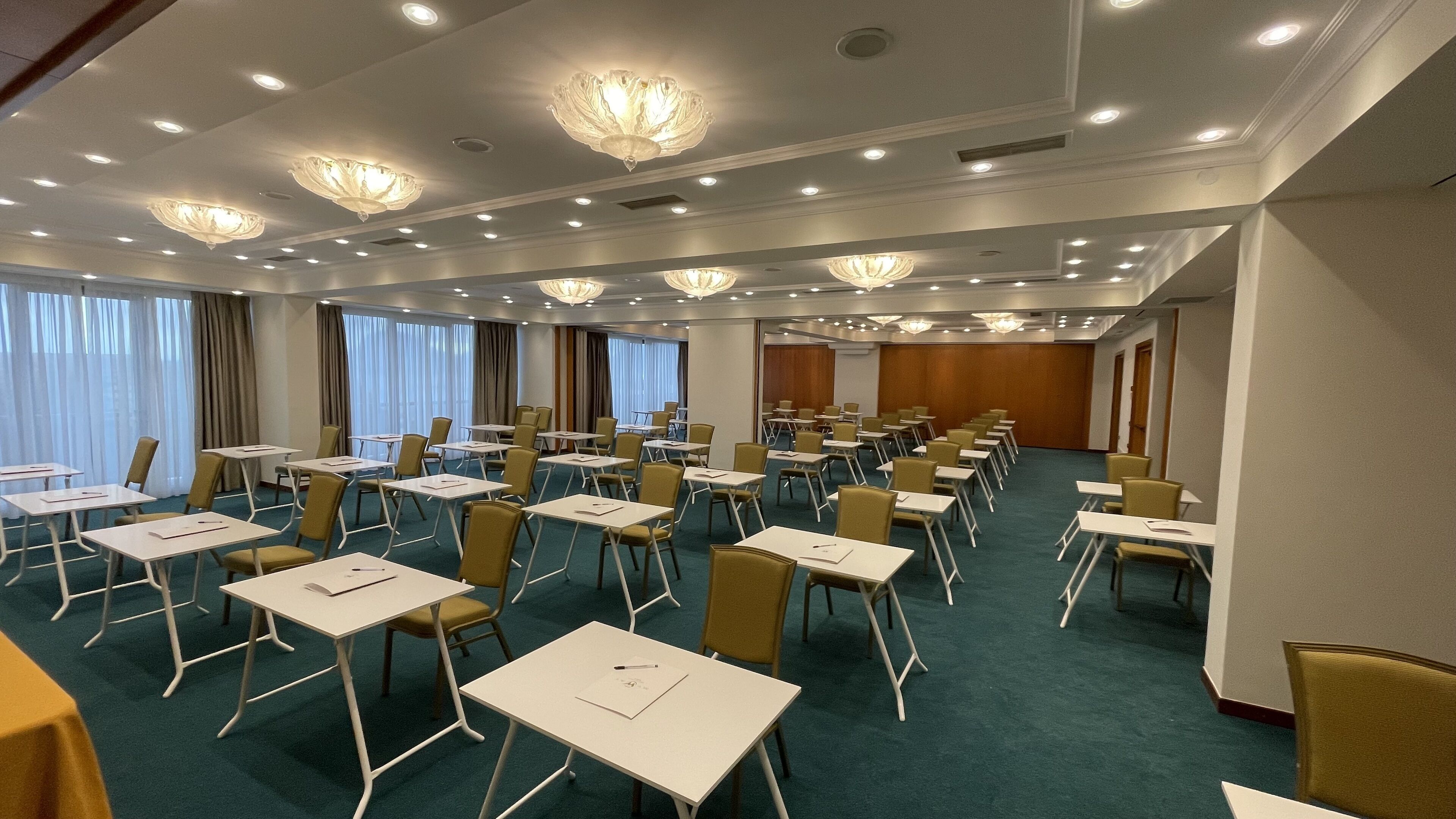 Un salón de conferencias con filas de mesas y sillas, ventanas amplias con cortinas e iluminación empotrada en el techo.