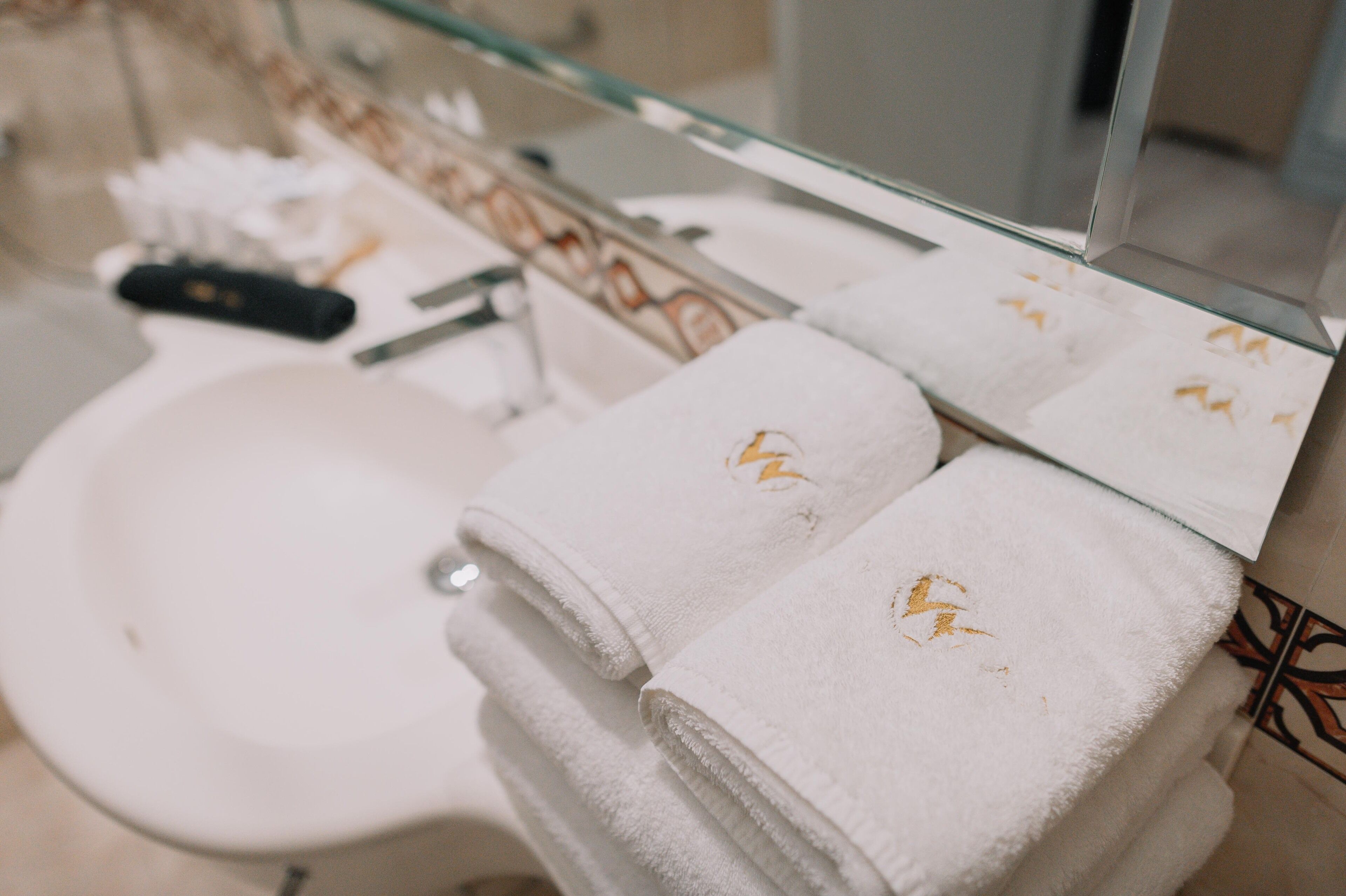 A bathroom with a white sink, a glass shower enclosure, and two towels with a gold emblem.