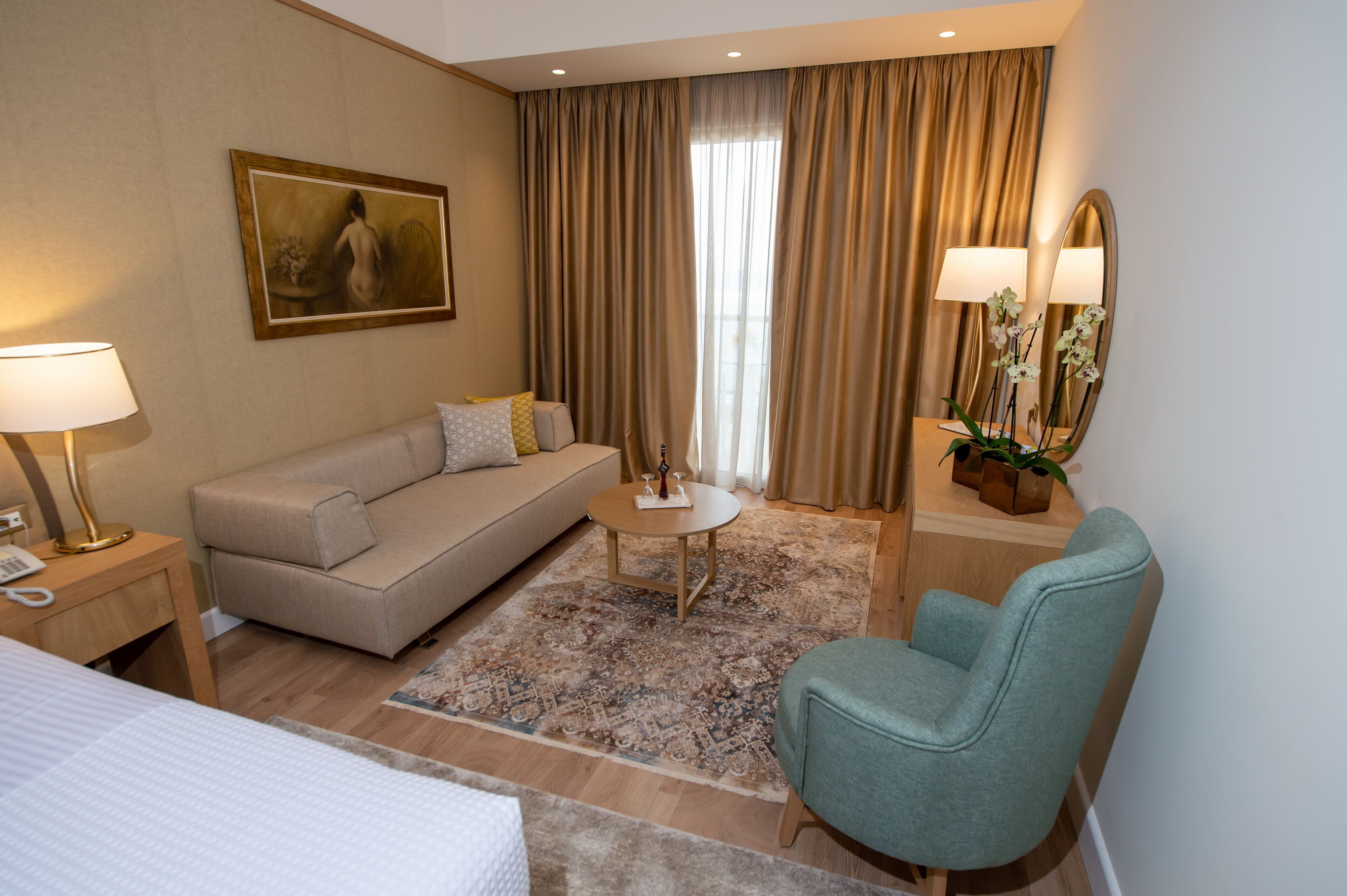 A hotel room with a sofa, a chair, a small table, a lamp, a mirror, and a painting on the wall.