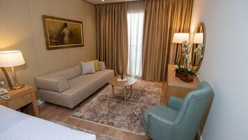 A hotel room with a sofa, a chair, a small table, a lamp, a mirror, and a painting on the wall.