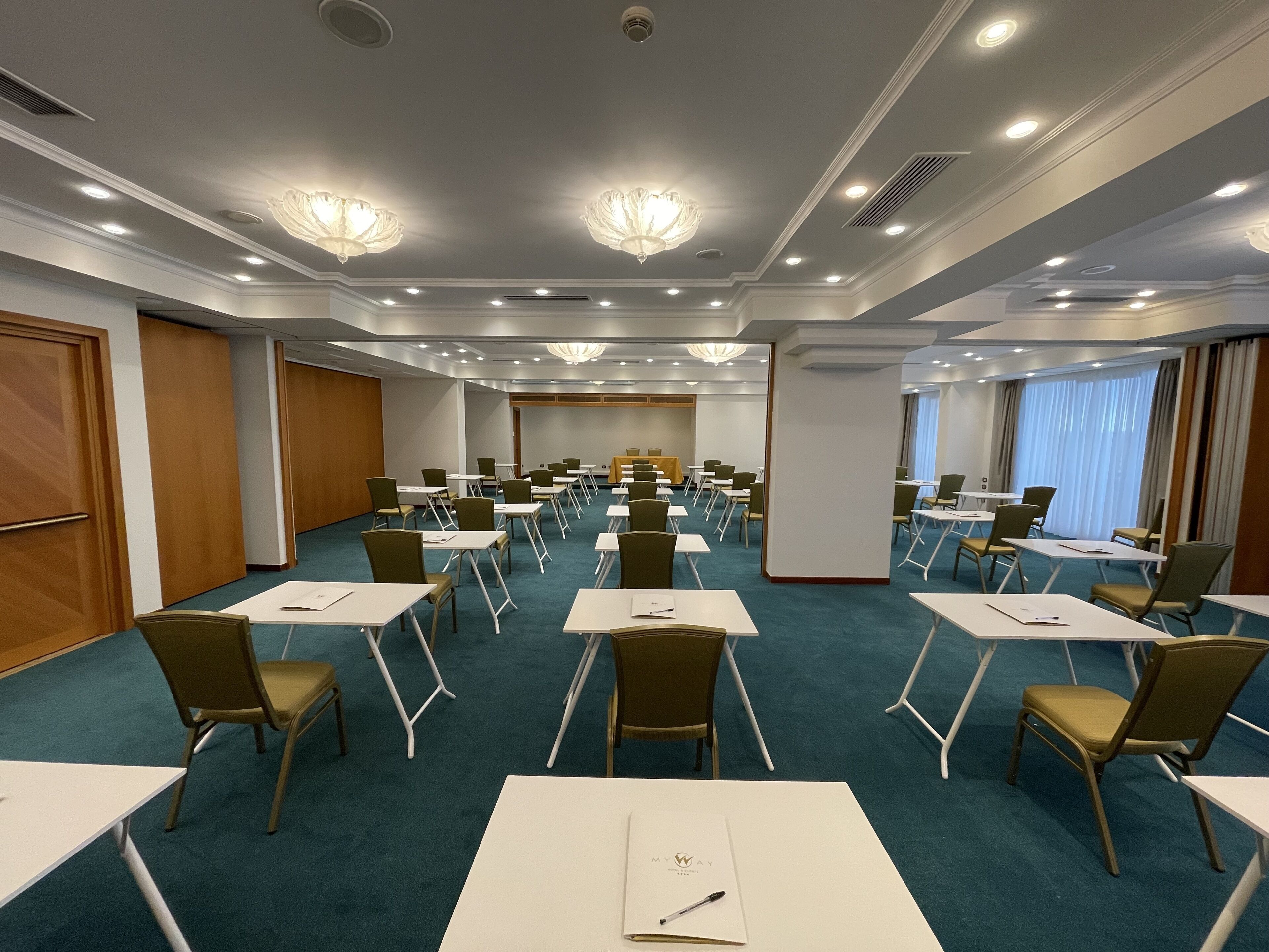 Un salón de conferencias con filas de mesas y sillas, iluminación empotrada y un candelabro.